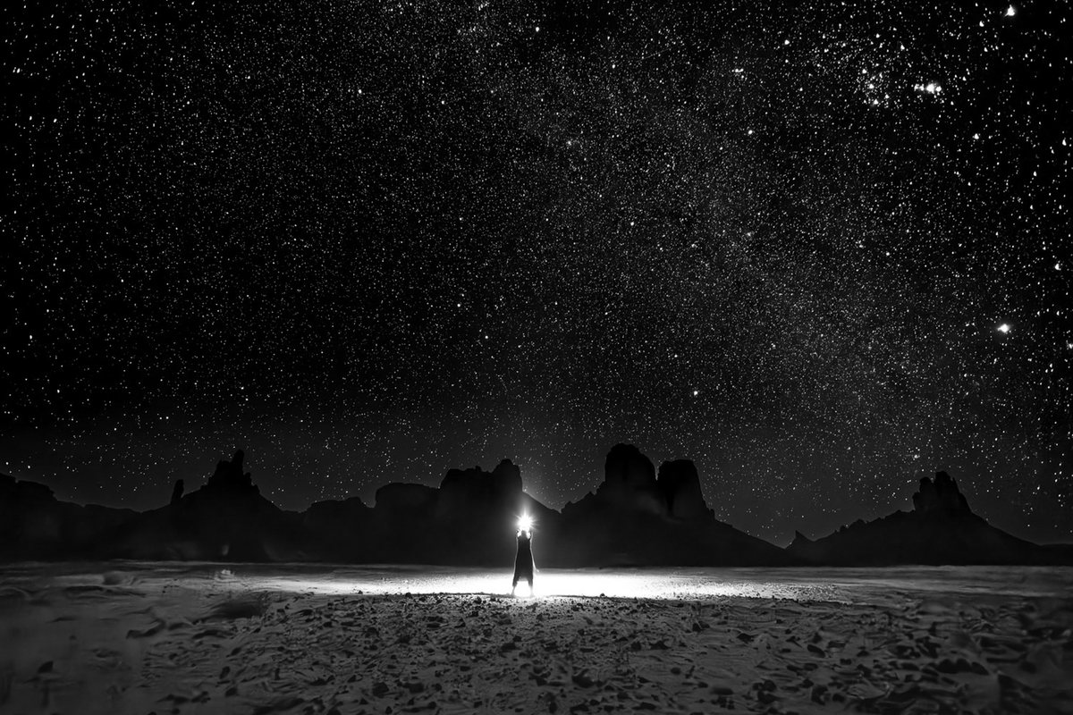 Un homme seul, les bras levés, tient une lumière au-dessus de sa tête, au milieu d’une plaine de nuit ; derrière lui, des silhouettes de rochers ; au-dessus, des milliers d’étoiles. Noir et blanc.