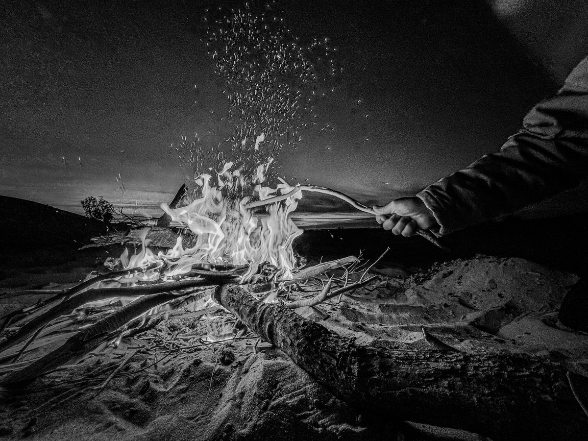 Une main attise un feu de bois posé sur le sable ; des étincelles s’élèvent dans la nuit. Noir et blanc.