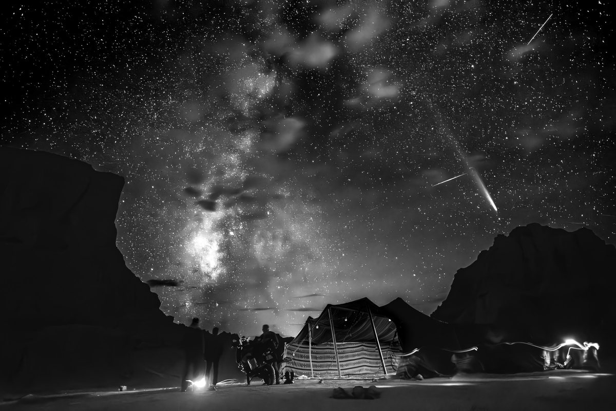 La Voie lactée et une étoile filante au-dessus d’un campement : une tente, quelques silhouettes, des traînées de lumière. Noir et blanc.