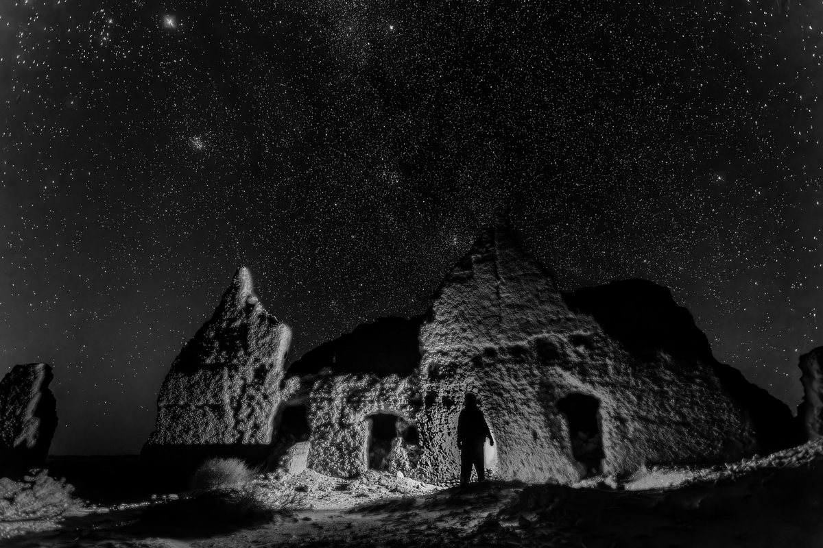 Un homme debout dans l’embrasure d’une ruine de terre éclairée, sous un ciel étoilé. Noir et blanc.