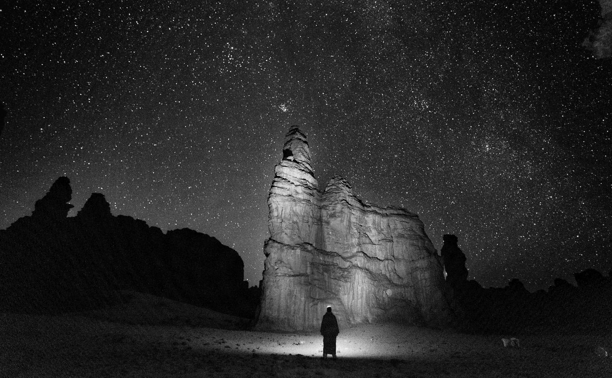Un homme de dos, lampe frontale allumée, au pied d’un grand rocher éclairé, sous un ciel couvert d’étoiles. Noir et blanc.